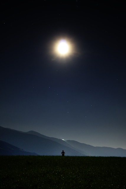 A bright moon shines down a person standing in a field, with hills in the background.